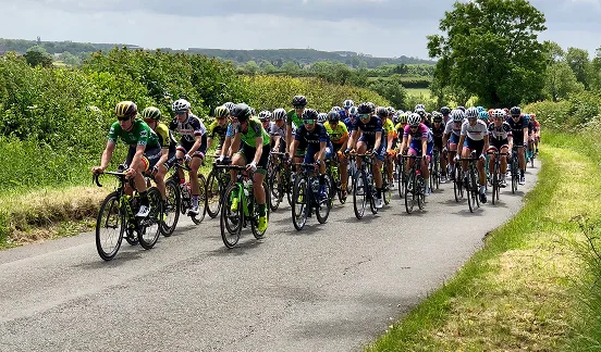 Un grand groupe de cyclistes vêtus de tenues colorées dévalent une route de campagne étroite et bordée d'arbres sous un ciel partiellement nuageux, dégageant une impression d'énergie et de compétition.
