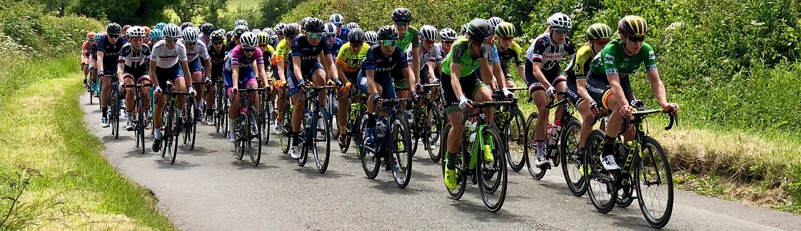 Un grand groupe de cyclistes vêtus de tenues colorées dévalent une route de campagne étroite et bordée d'arbres sous un ciel partiellement nuageux, dégageant une impression d'énergie et de compétition.