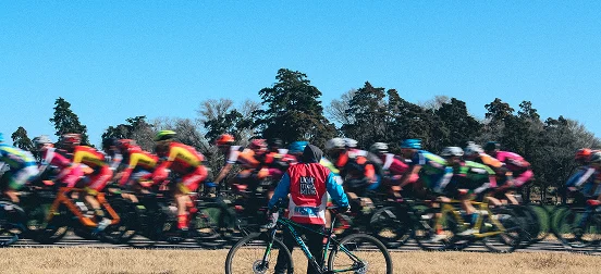 Un officiel de course vêtu d'un gilet rouge tient un vélo et observe un groupe flou de cyclistes qui passent à toute vitesse sous un ciel bleu limpide.