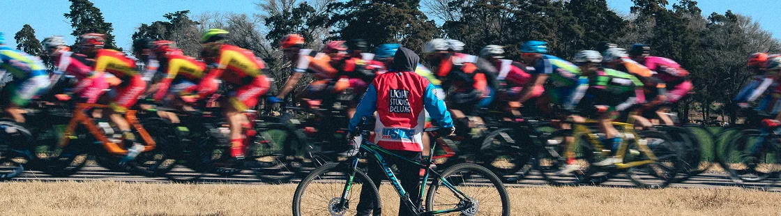 Un officiel de course vêtu d'un gilet rouge tient un vélo et observe un groupe flou de cyclistes qui passent à toute vitesse sous un ciel bleu limpide.